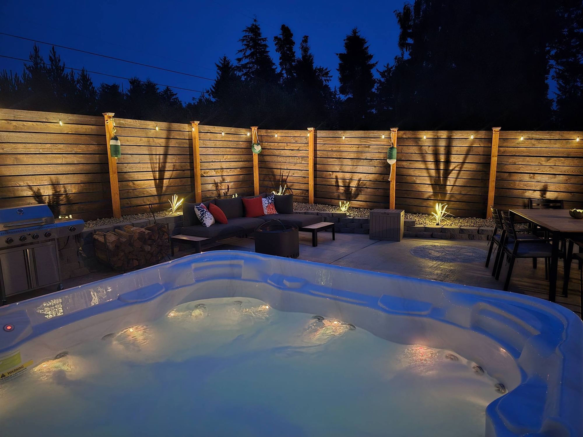The lit hot tub at blue hour, with the fire pit lounge, grill and dining table inside the cedar fence